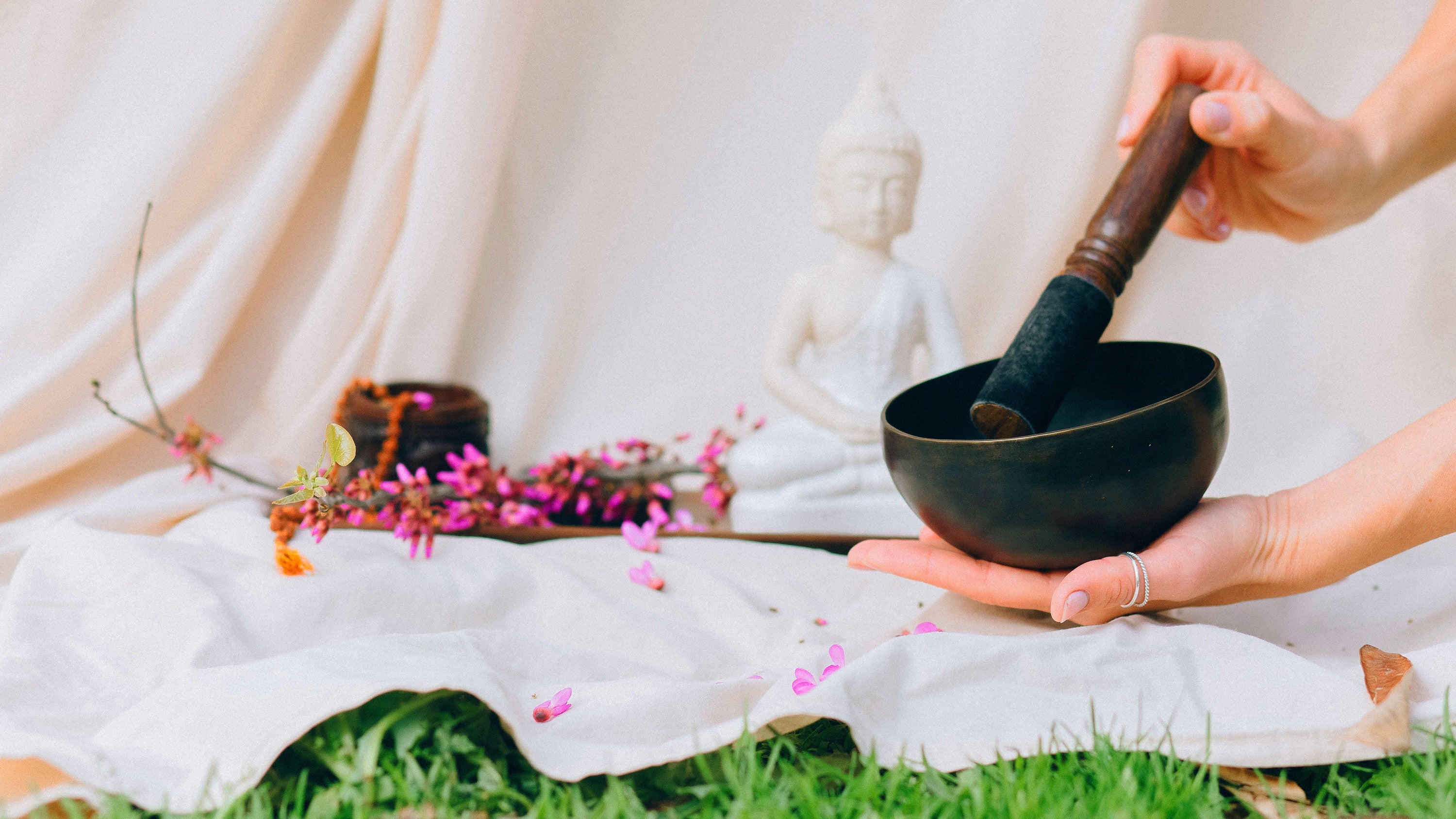 A close shot of a sound bowl being played with a mallet with a buddha statue and bed sheet in the background.